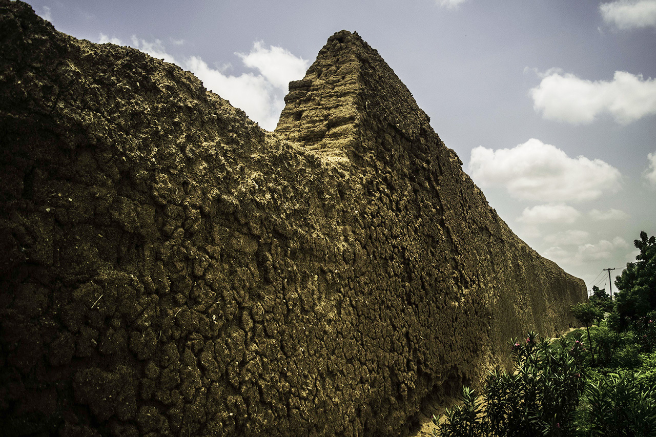 A section of the old city wall of Kano, a rough earthen rampart under a partly cloudy sky