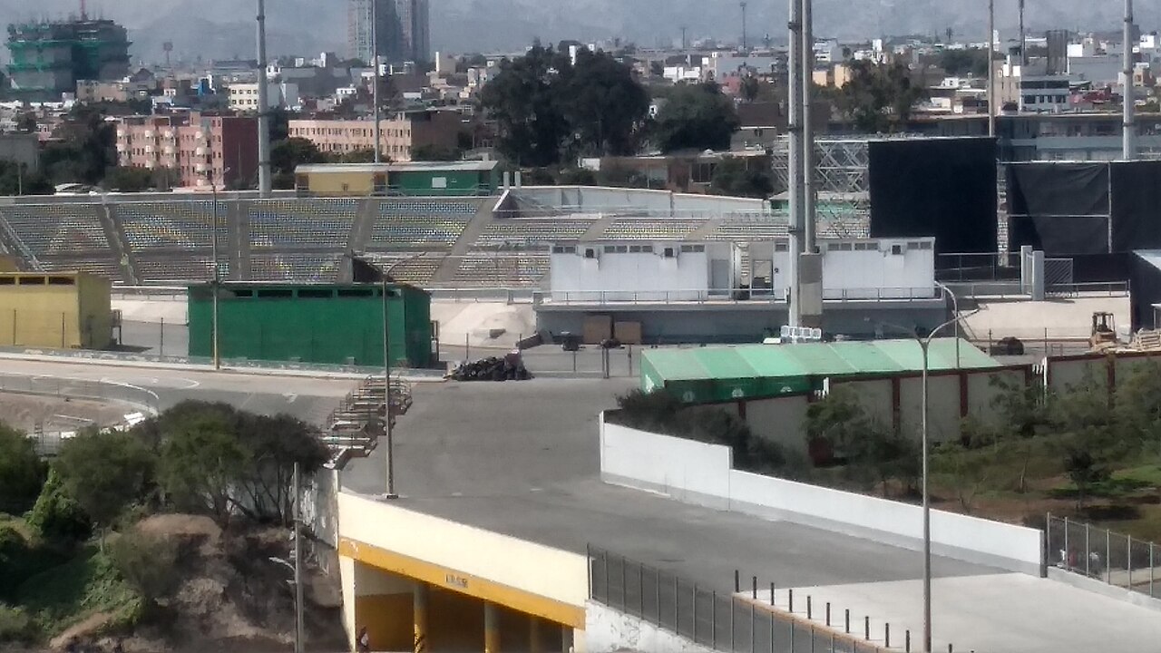 The Estadio San Marcos in Lima seen from outside, with empty stands, temporary buildings and black-draped scaffolding, and city buildings and hills behind.