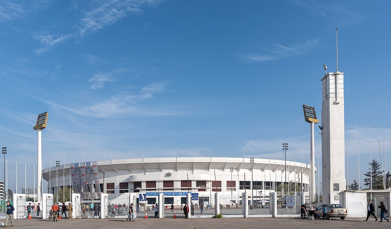 The white oval stand of Santiago's Estadio Nacional behind a fenced entrance plaza, with floodlight towers, a white tower and people walking under a blue sky.