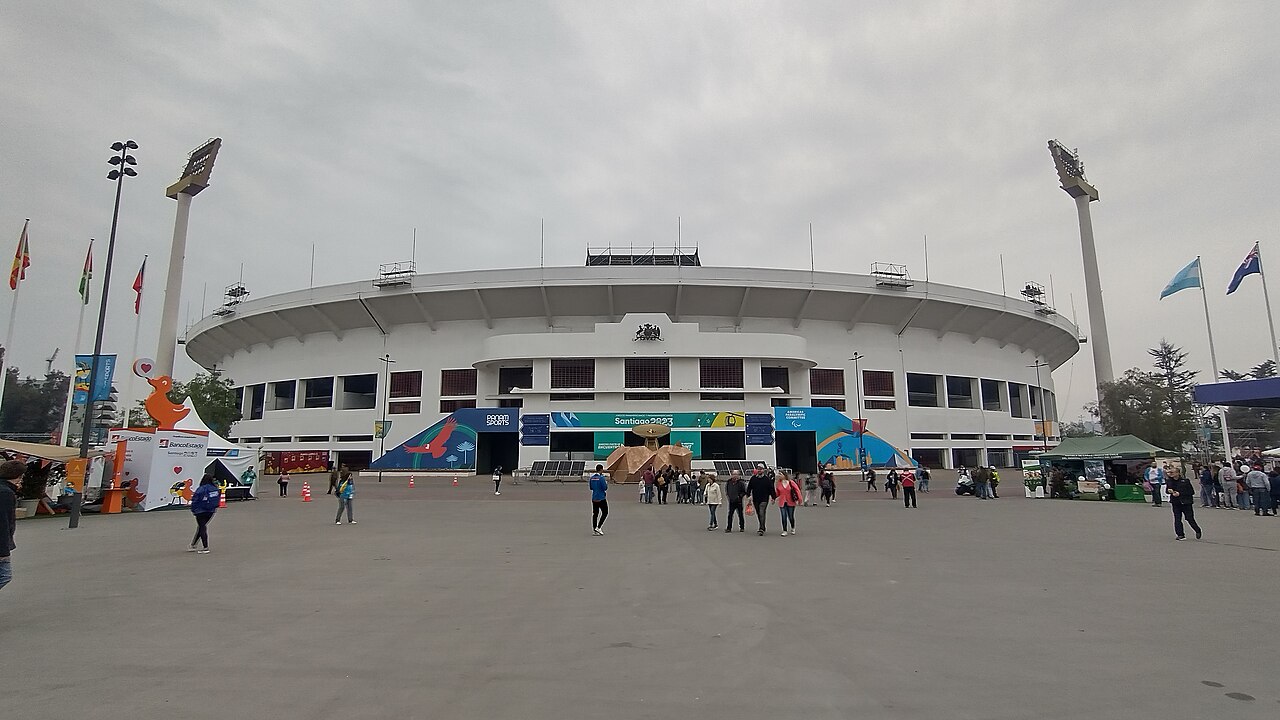 Fans walk across a wide concrete plaza towards the main facade of Santiago's Estadio Nacional, with flags and floodlights under a grey sky.