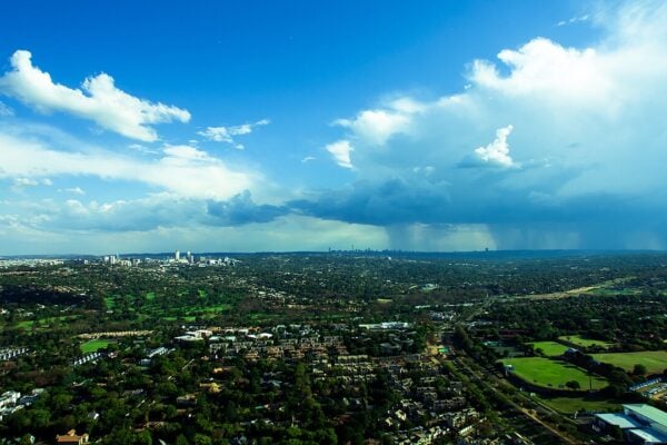 Aerial view of tree-lined suburbs of Johannesburg under a blue sky with a rain storm and the Sandton skyline in the distance