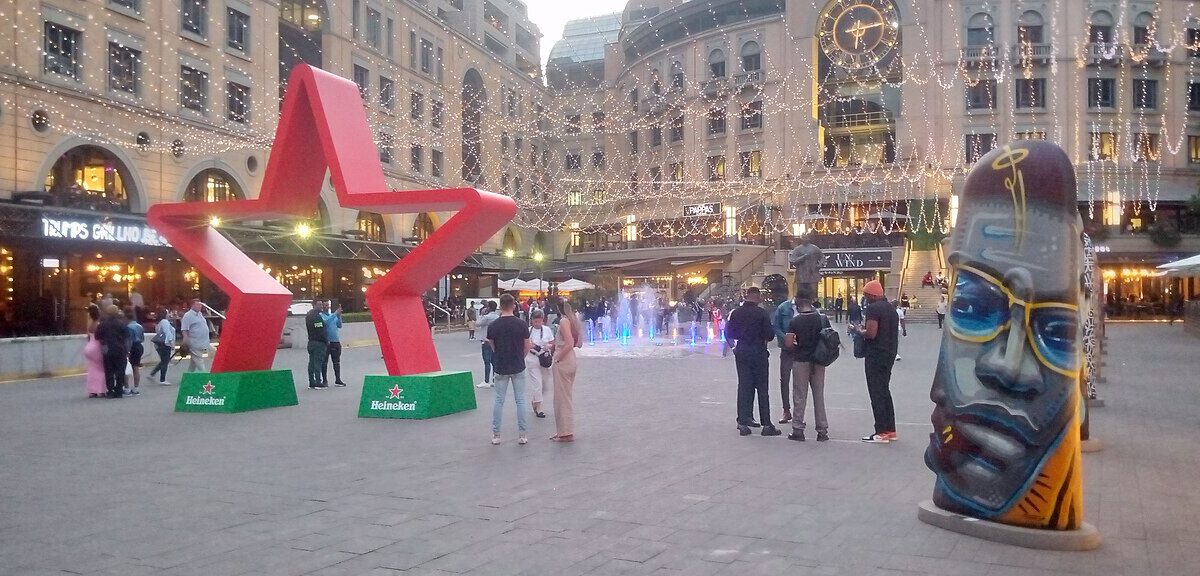 Shoppers and visitors on Nelson Mandela Square in Sandton, Johannesburg, with a large red star sculpture and a painted head sculpture