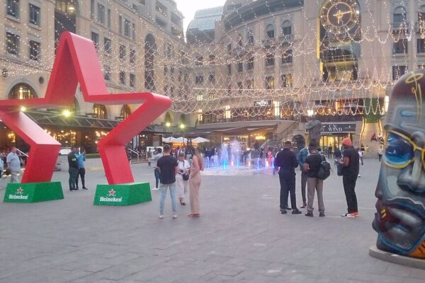 Shoppers and visitors on Nelson Mandela Square in Sandton, Johannesburg, with a large red star sculpture and a painted head sculpture