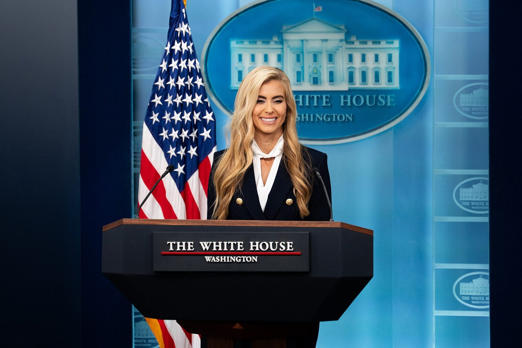 Press Secretary Katie Zacharia at the lectern of the White House briefing room, smiling, with the American flag behind her