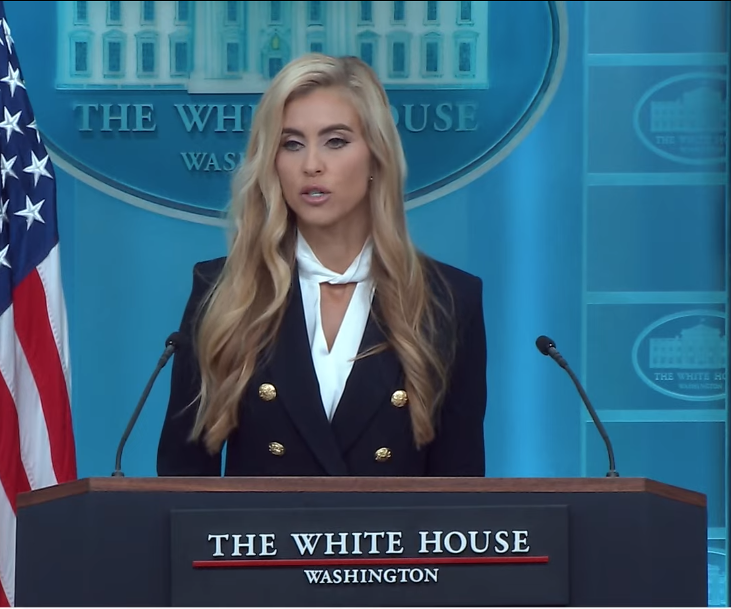Katie Zacharia speaking at the White House briefing room lectern, in a dark double-breasted blazer