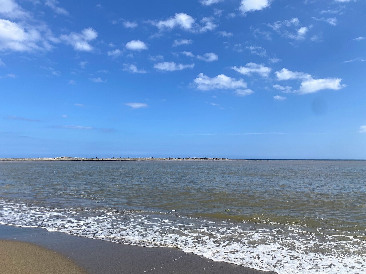 Sandy shore and calm brown-green sea under a blue sky with small clouds, a low breakwater on the horizon, Playa Montesinos, Santo Domingo