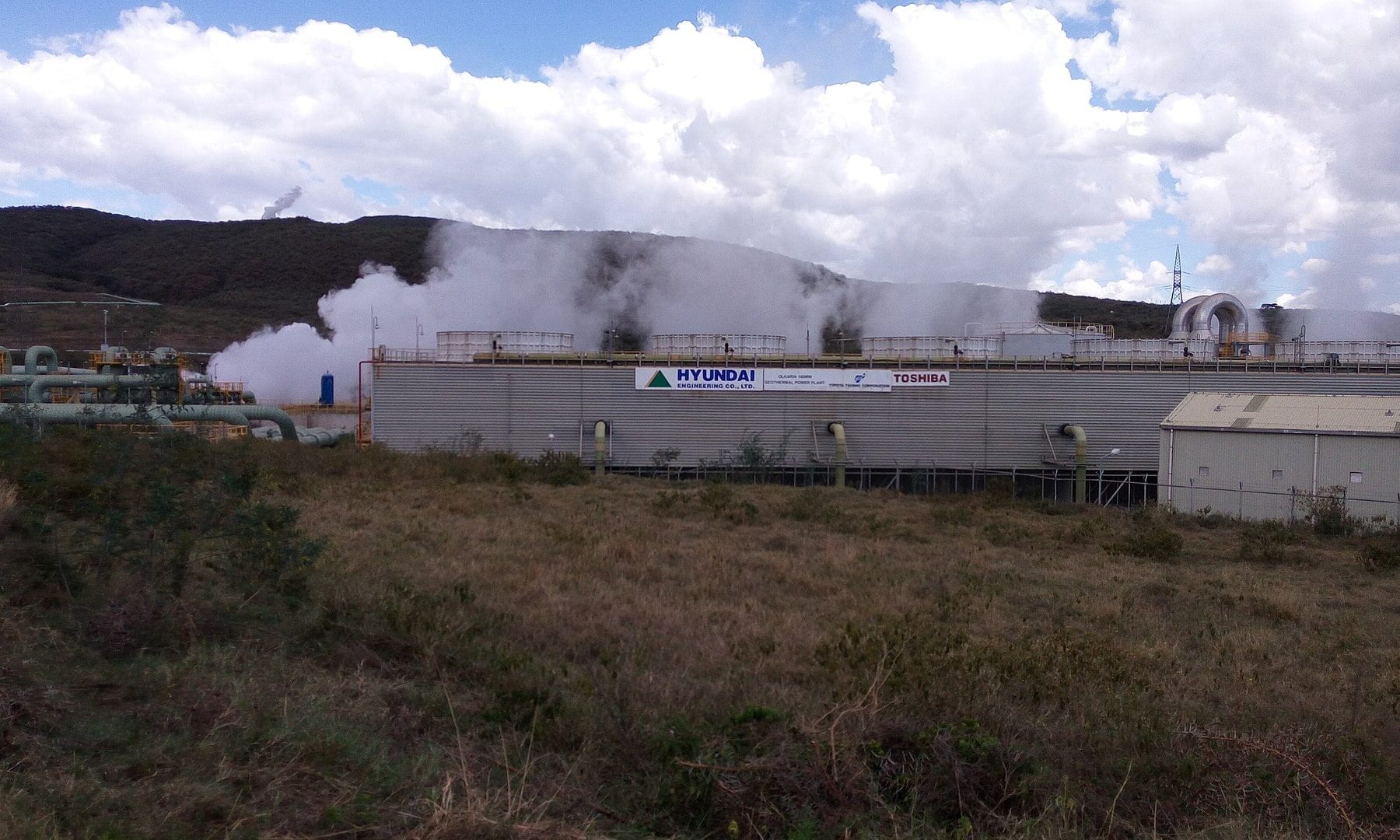 White steam rises from the cooling towers of a geothermal power plant at Olkaria, Kenya, with pipes in the foreground and a hill behind