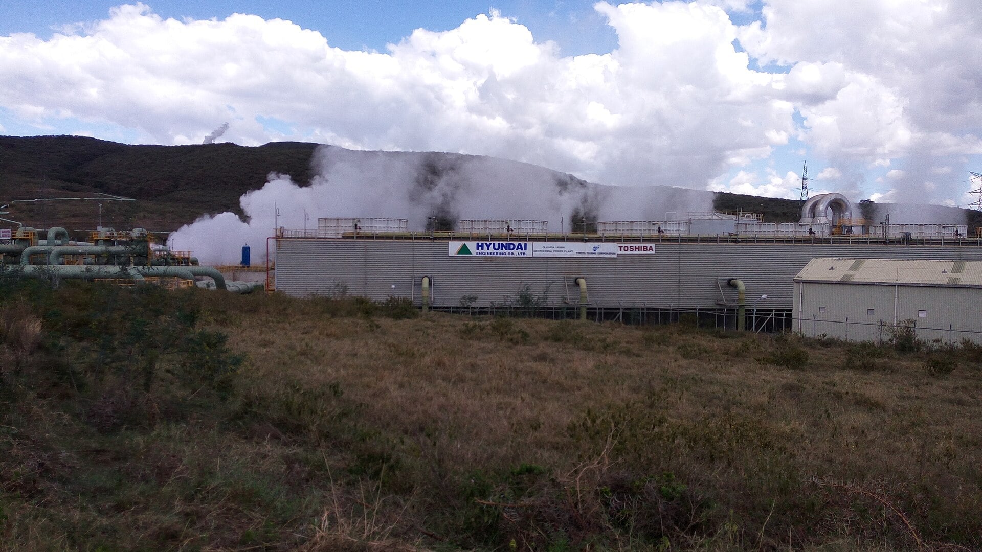 White steam rises from the cooling towers of a geothermal power plant at Olkaria, Kenya, with pipes in the foreground and a hill behind