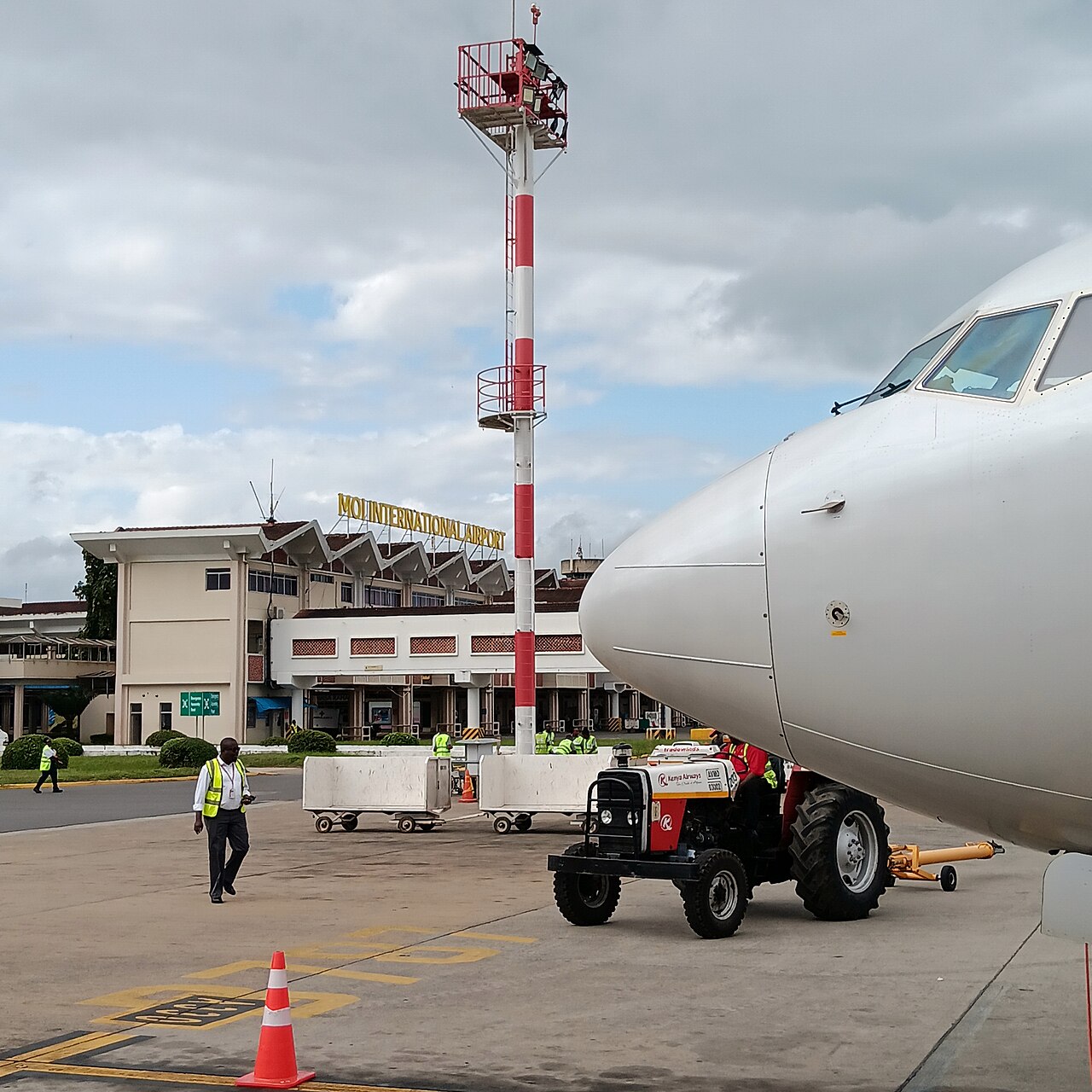 Kenya Airways ground crew with a baggage tractor and carts beside the nose of a Boeing 737 at Moi International Airport