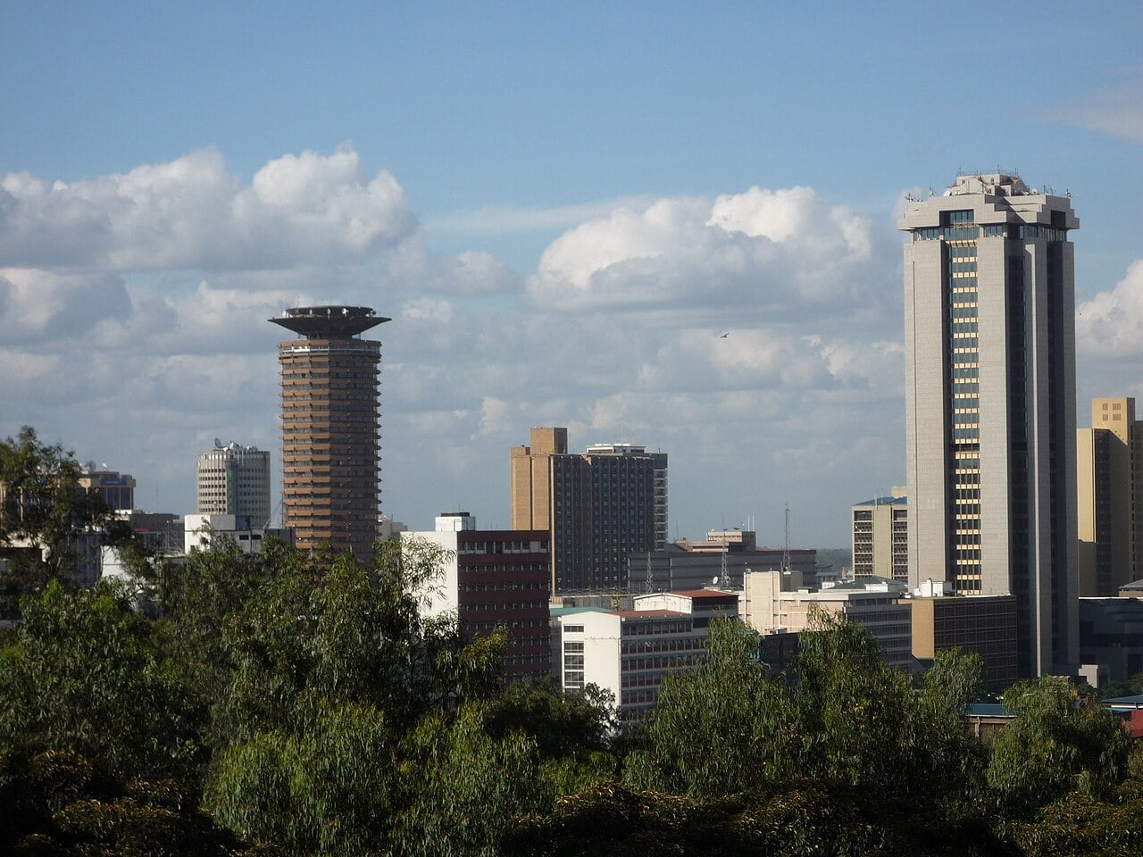 Office towers in central Nairobi under a blue sky with white clouds, seen over treetops