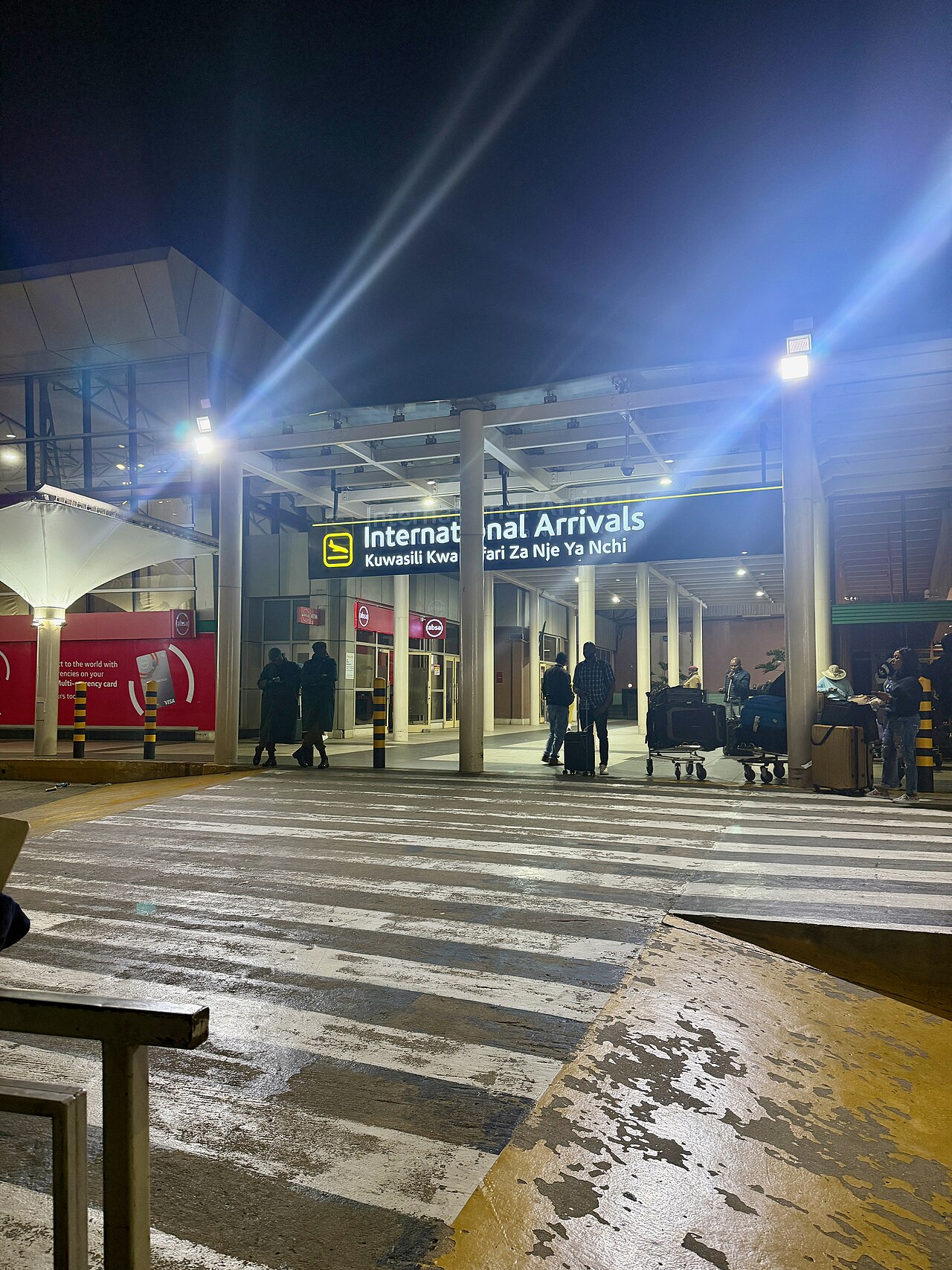 International Arrivals sign in English and Swahili at Jomo Kenyatta International Airport at night, with travellers and luggage