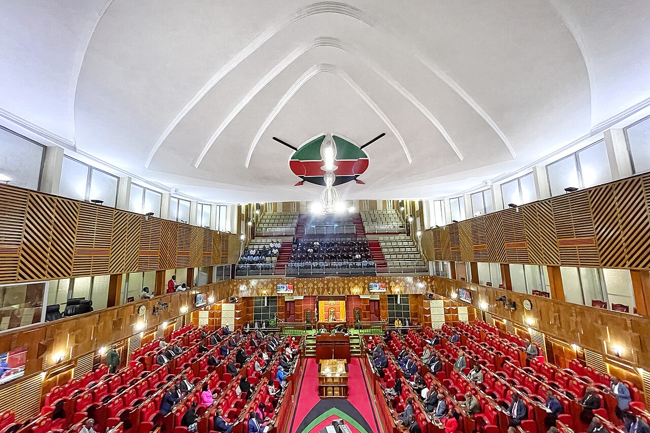 Debating chamber of Kenya's National Assembly in Nairobi, seen from the gallery, with red benches, wooden panelling and a shield emblem on the ceiling
