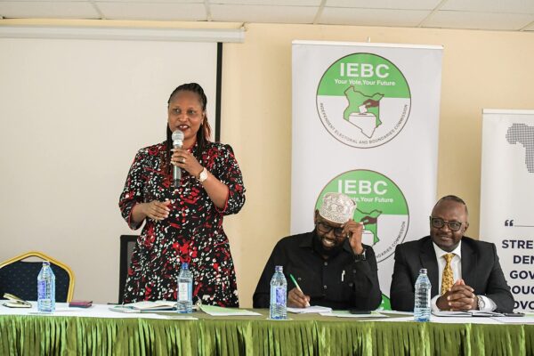 Woman speaking at a microphone beside two seated men in front of banners of the Independent Electoral and Boundaries Commission in Kenya