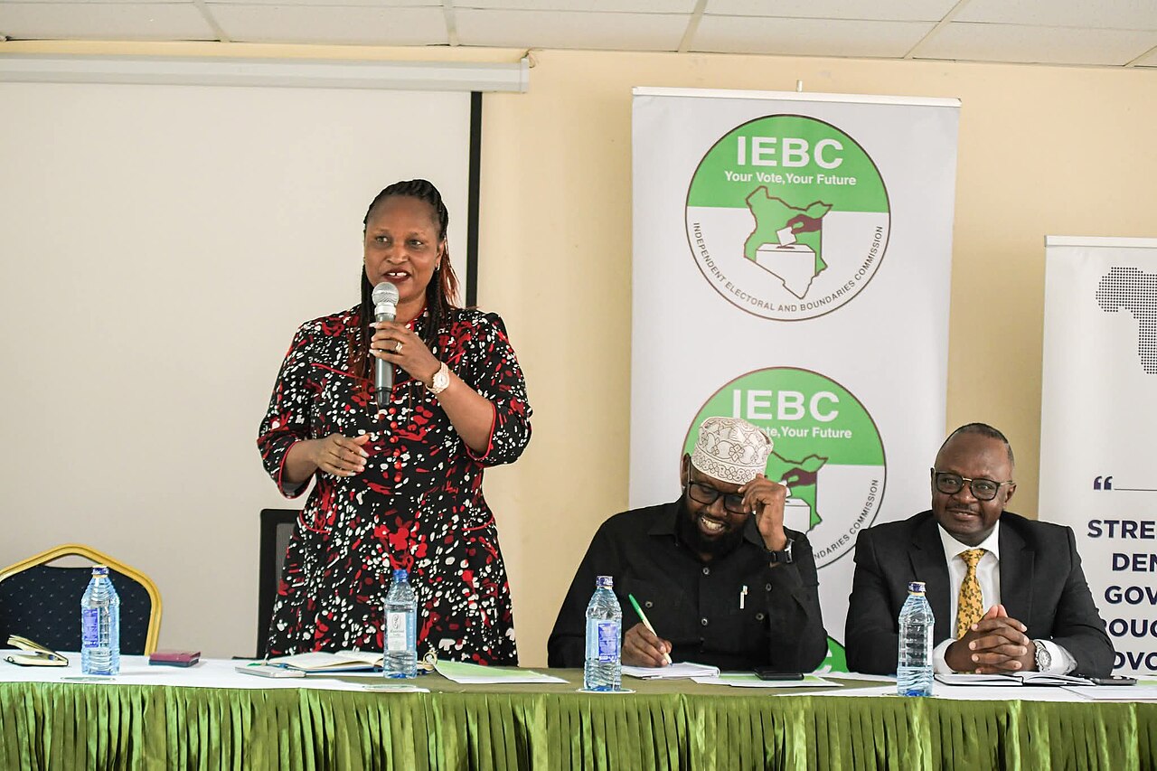 Woman speaking at a microphone beside two seated men in front of banners of the Independent Electoral and Boundaries Commission in Kenya