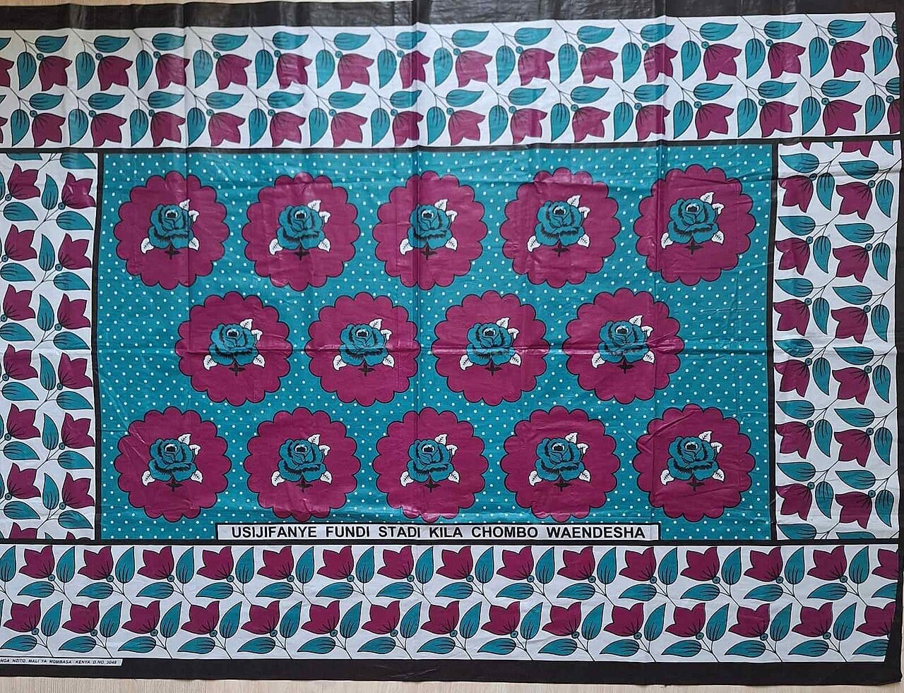 A Kenyan kanga cloth printed with magenta roses and teal leaves on a polka-dot field with a Swahili line along the bottom