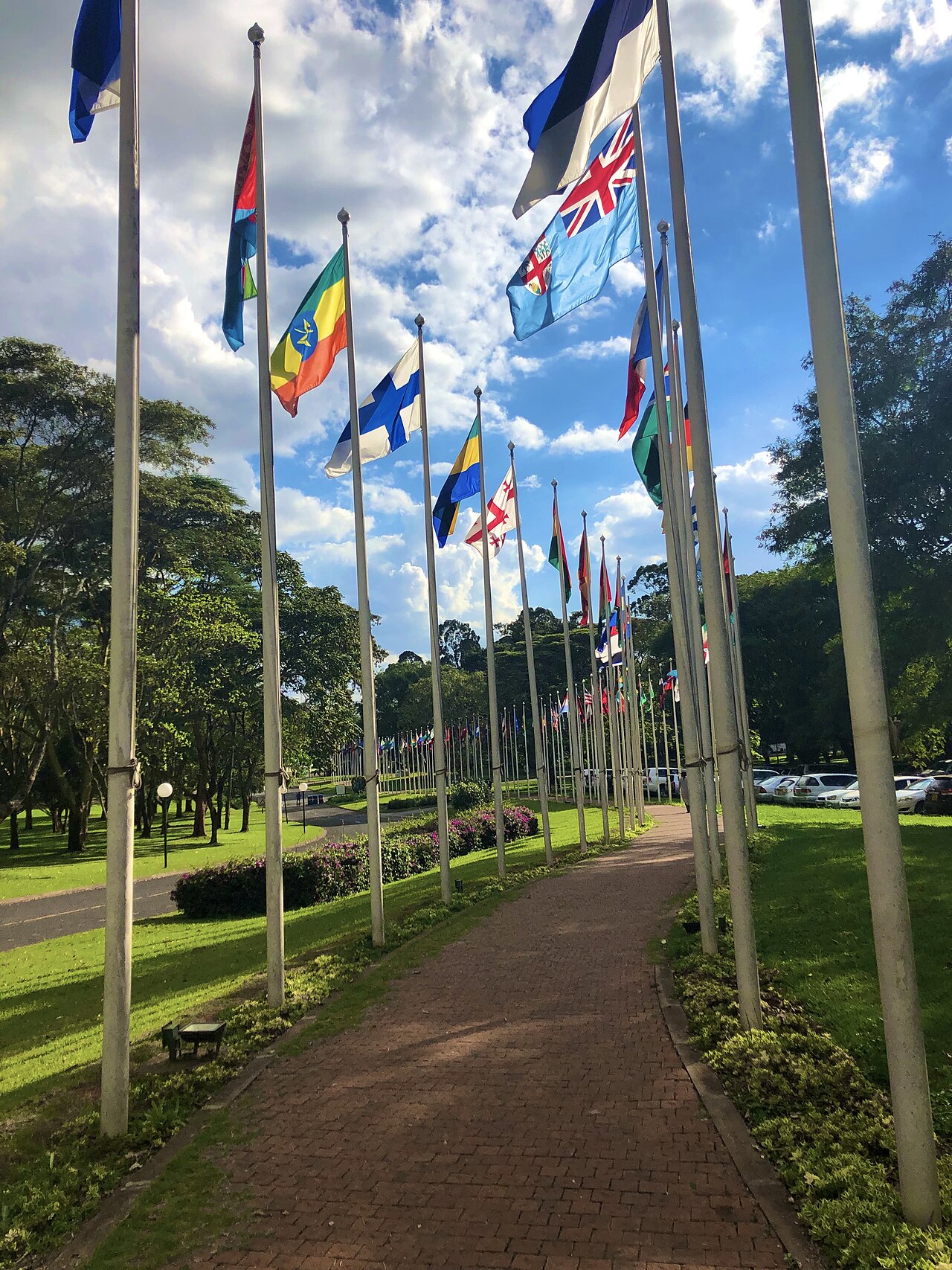 Rows of national flags on tall poles lining a brick footpath through green lawns at the United Nations Office at Nairobi