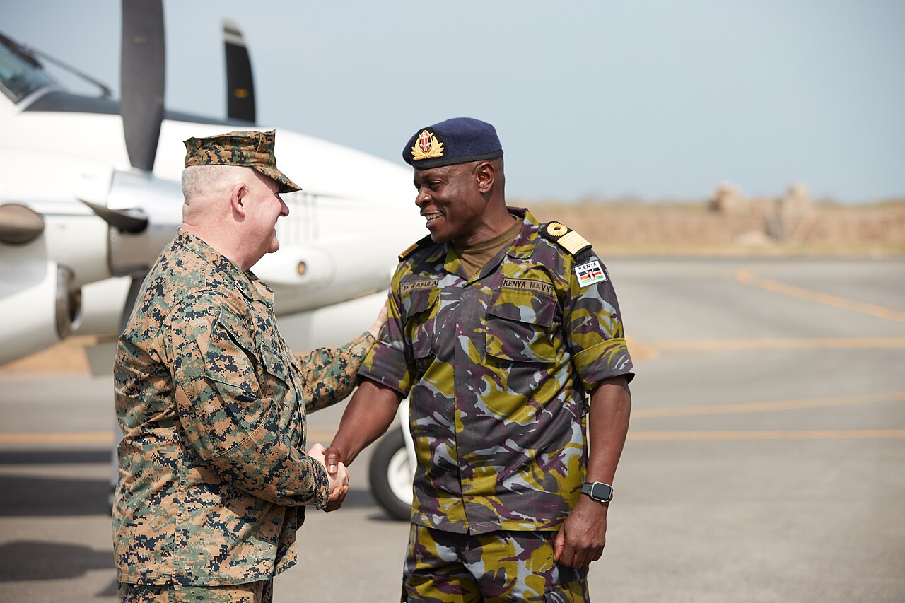 A US Marine Corps general in camouflage shakes hands with a smiling Kenya Navy officer in a blue beret beside a propeller aircraft on the Manda Bay airfield