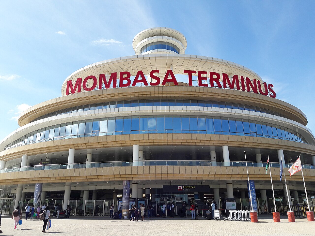 Curved glass and cream facade of Mombasa Terminus railway station with red lettering and travellers walking to the entrance