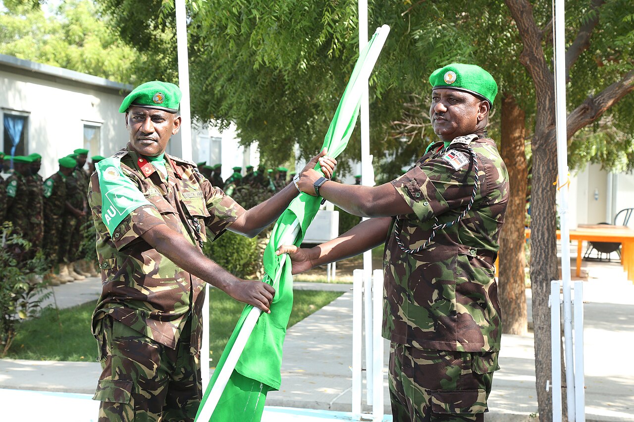 Two Kenyan officers in camouflage and green berets hold a green African Union flag as soldiers stand in formation behind them