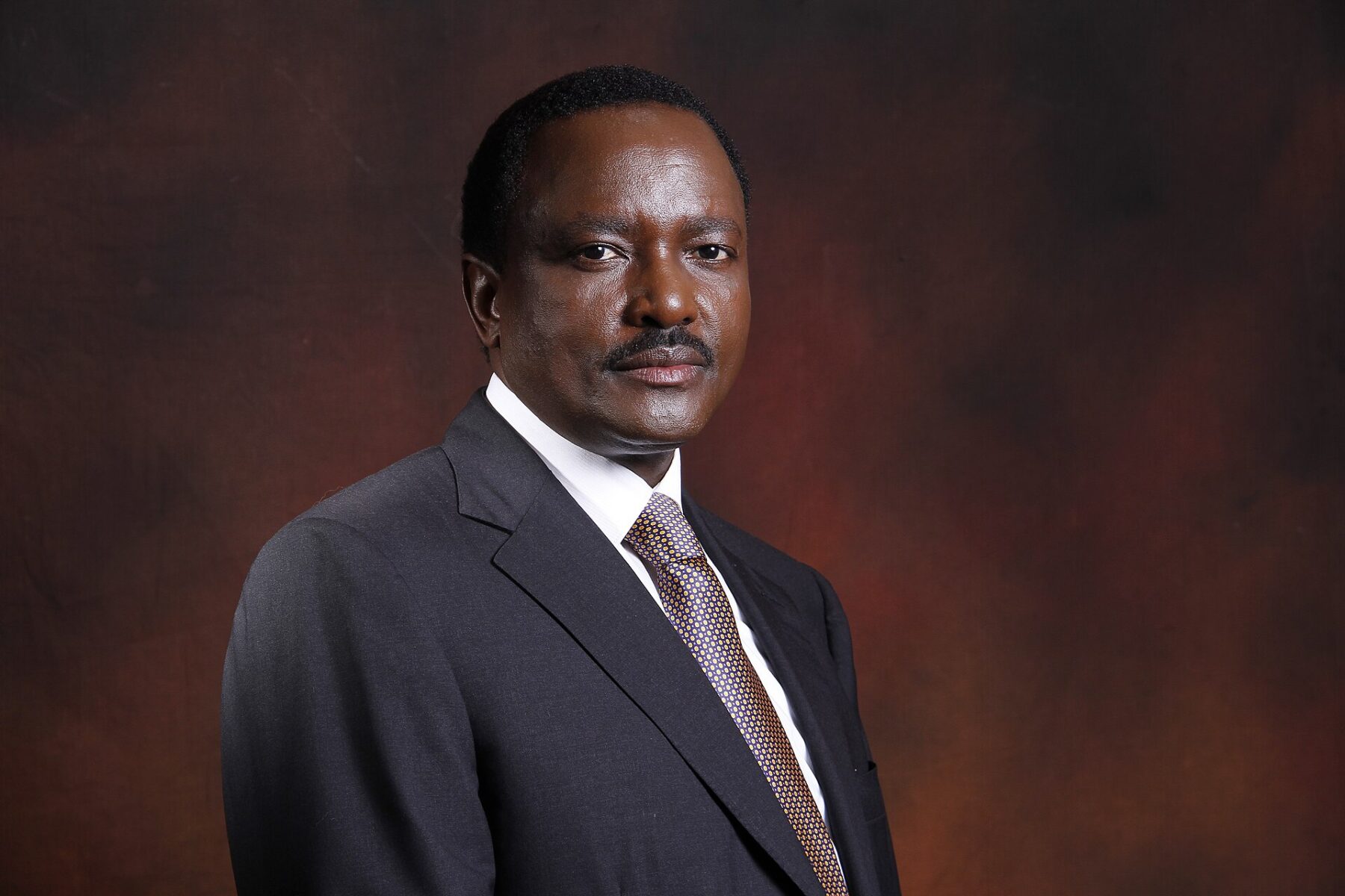 Kalonzo Musyoka, a Kenyan politician in a dark suit and tie, looks at the camera against a dark brown studio background