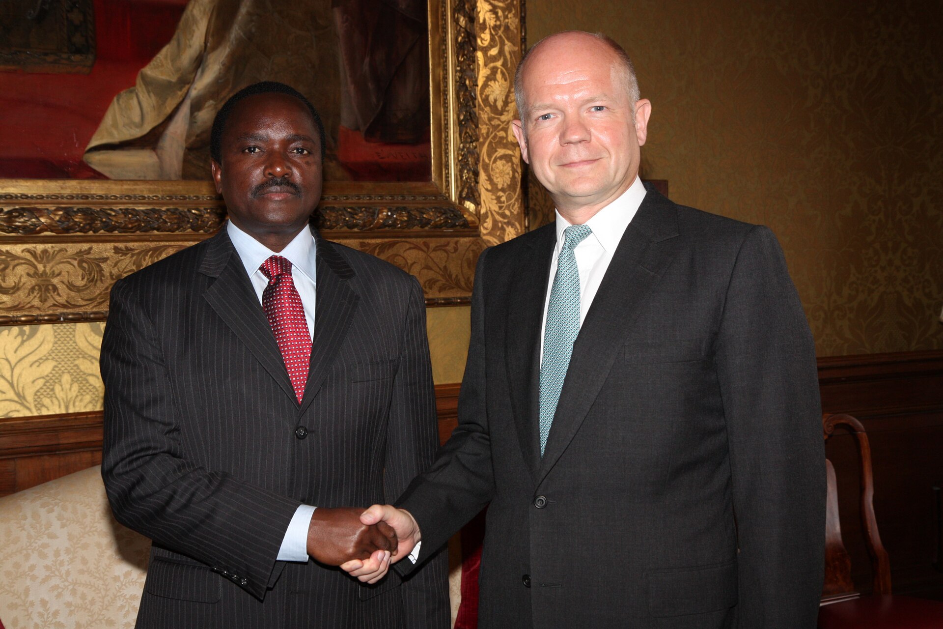 Kalonzo Musyoka and British Foreign Secretary William Hague shake hands in front of a gilt-framed painting in London