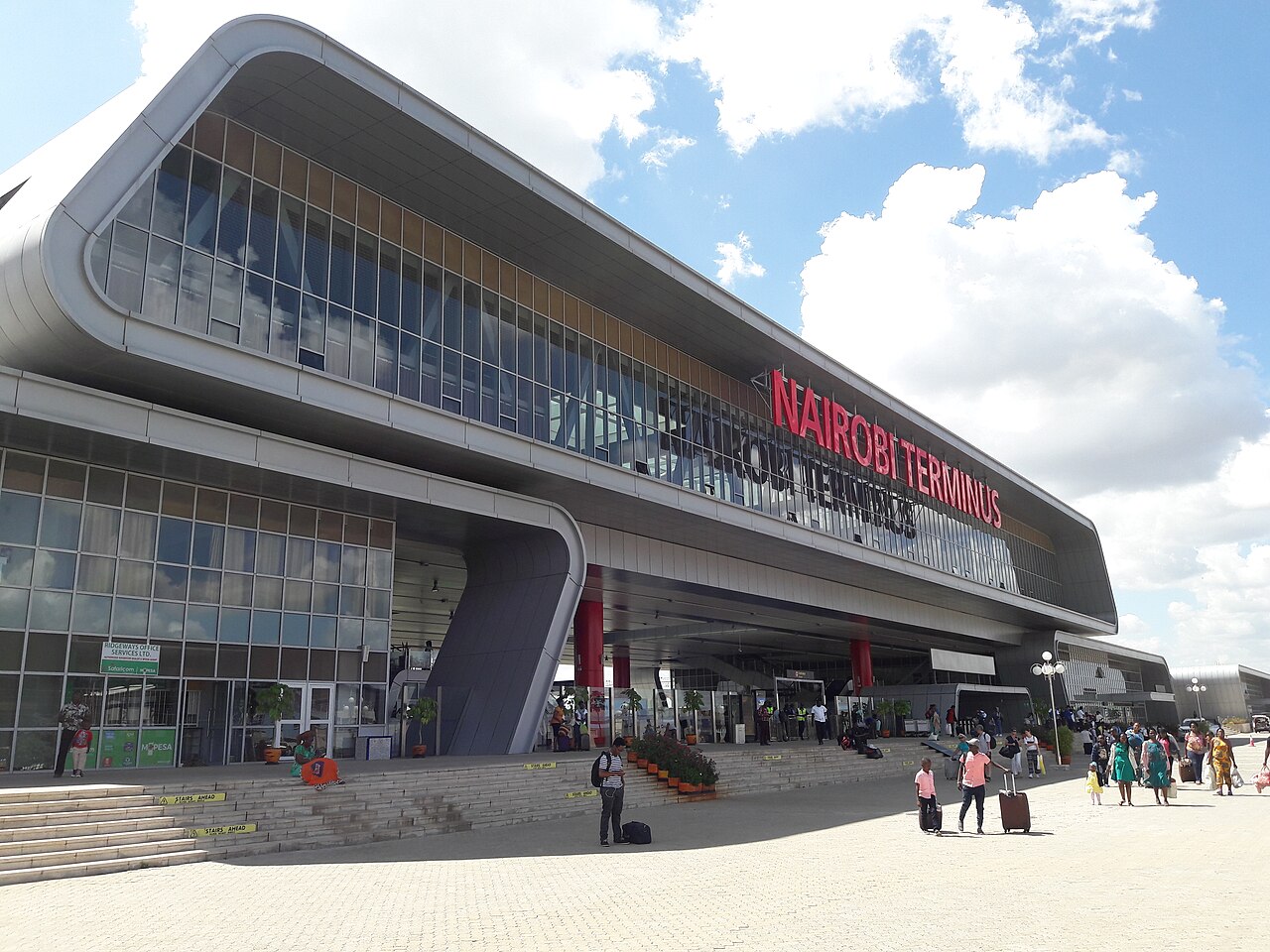 Entrance of Nairobi Terminus railway station with its curved glass and steel facade, and travellers with luggage in front under a blue sky