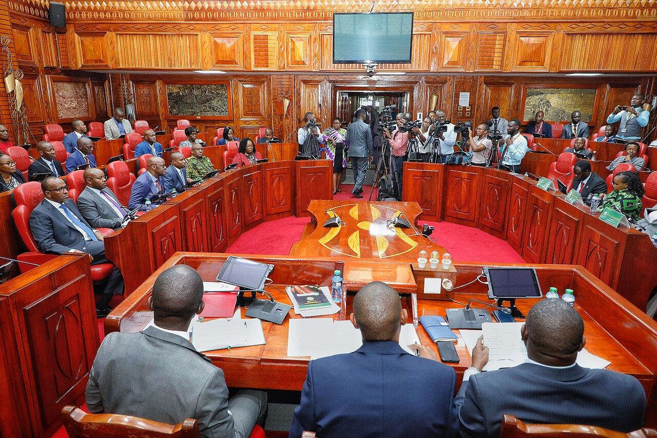 Journalists with cameras on tripods film a committee meeting in a wood-panelled room of Kenya's National Assembly, with red leather seats