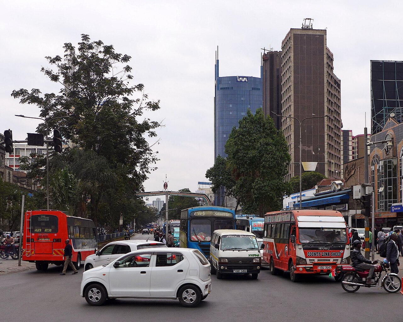 Red and blue buses, a white minibus, cars and a motorbike on Kenyatta Avenue in central Nairobi, Kenya, with office towers behind
