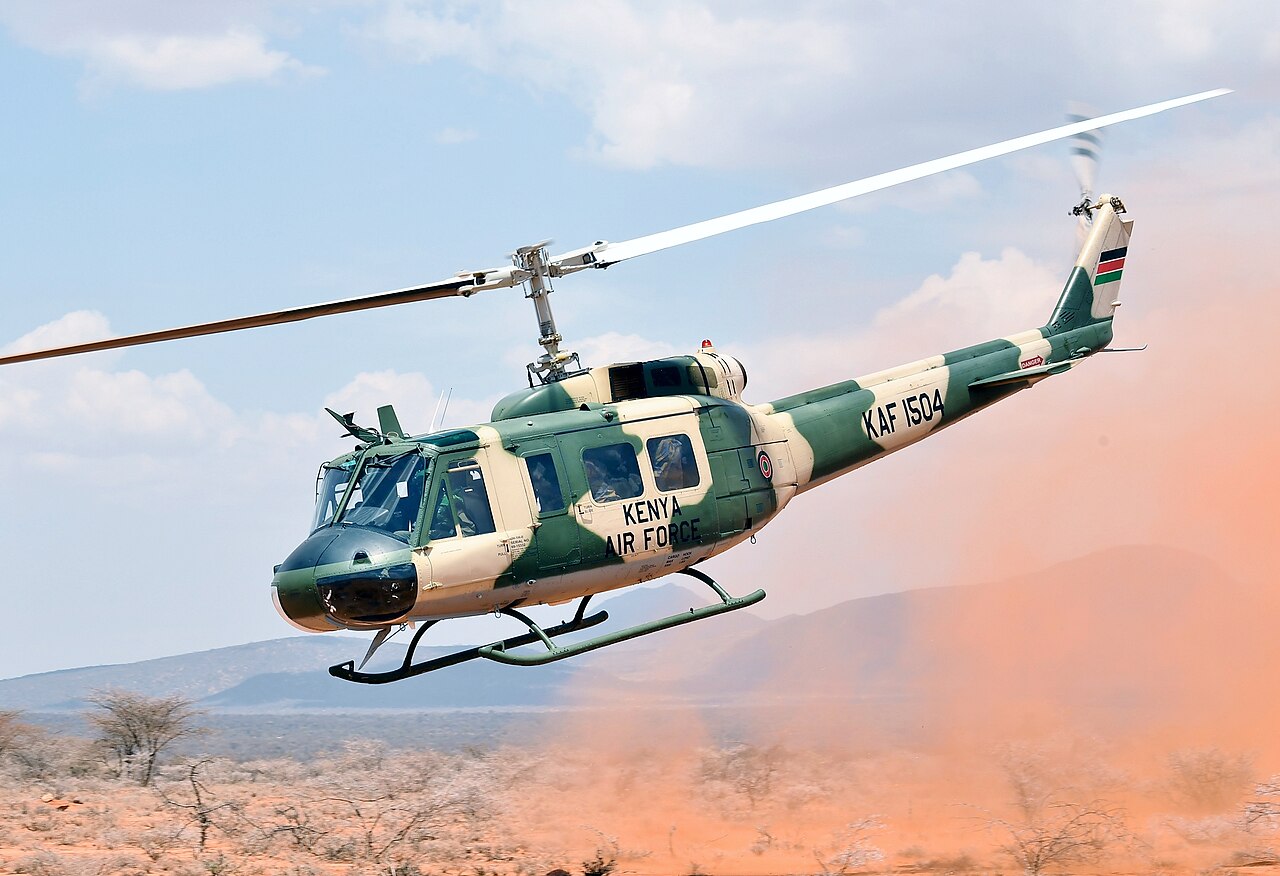 A green and cream Kenya Air Force UH-1 Huey helicopter, tail number KAF 1504, lifts off in a cloud of red dust with hills behind