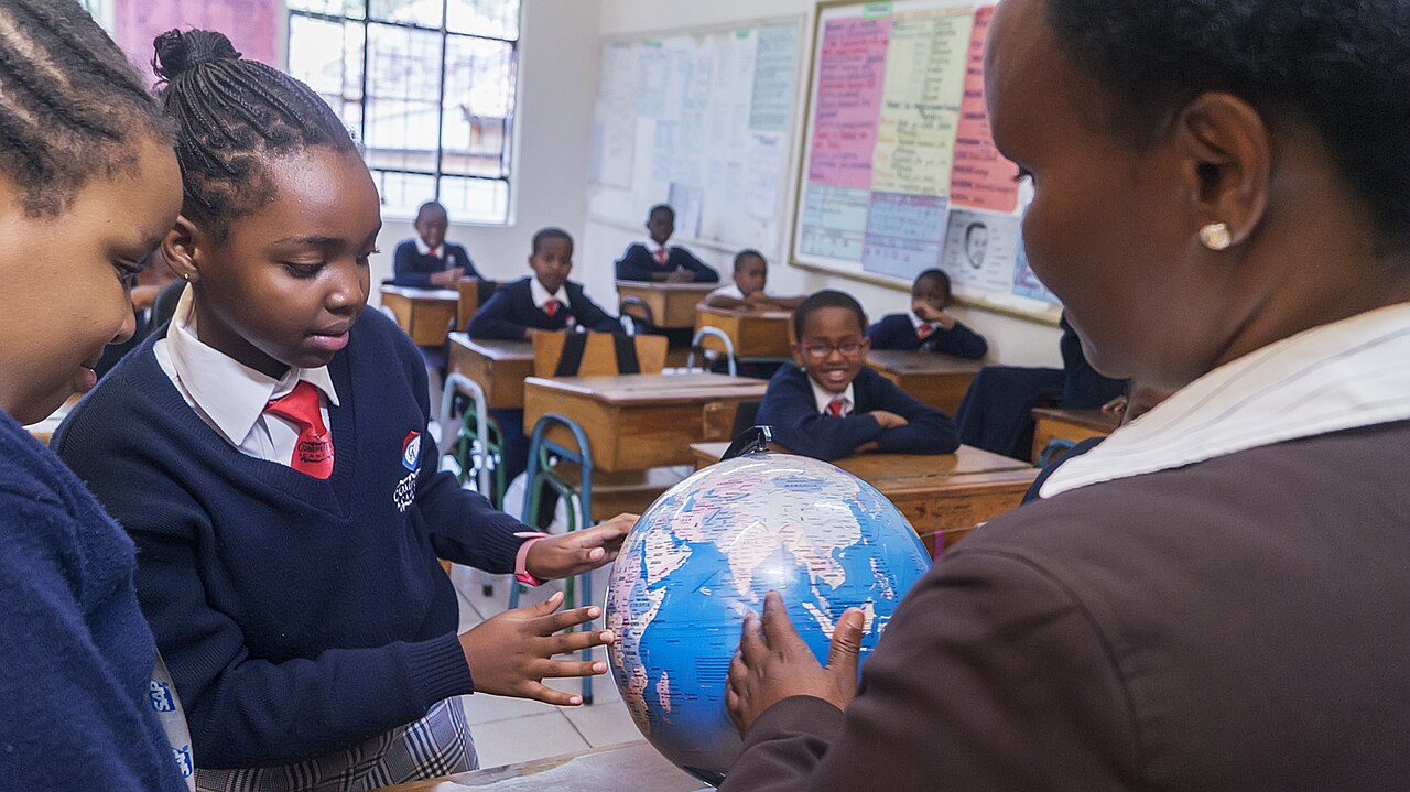 A teacher holds a globe as two girls in navy school uniforms look on in a Kenyan classroom, with pupils seated at wooden desks behind them