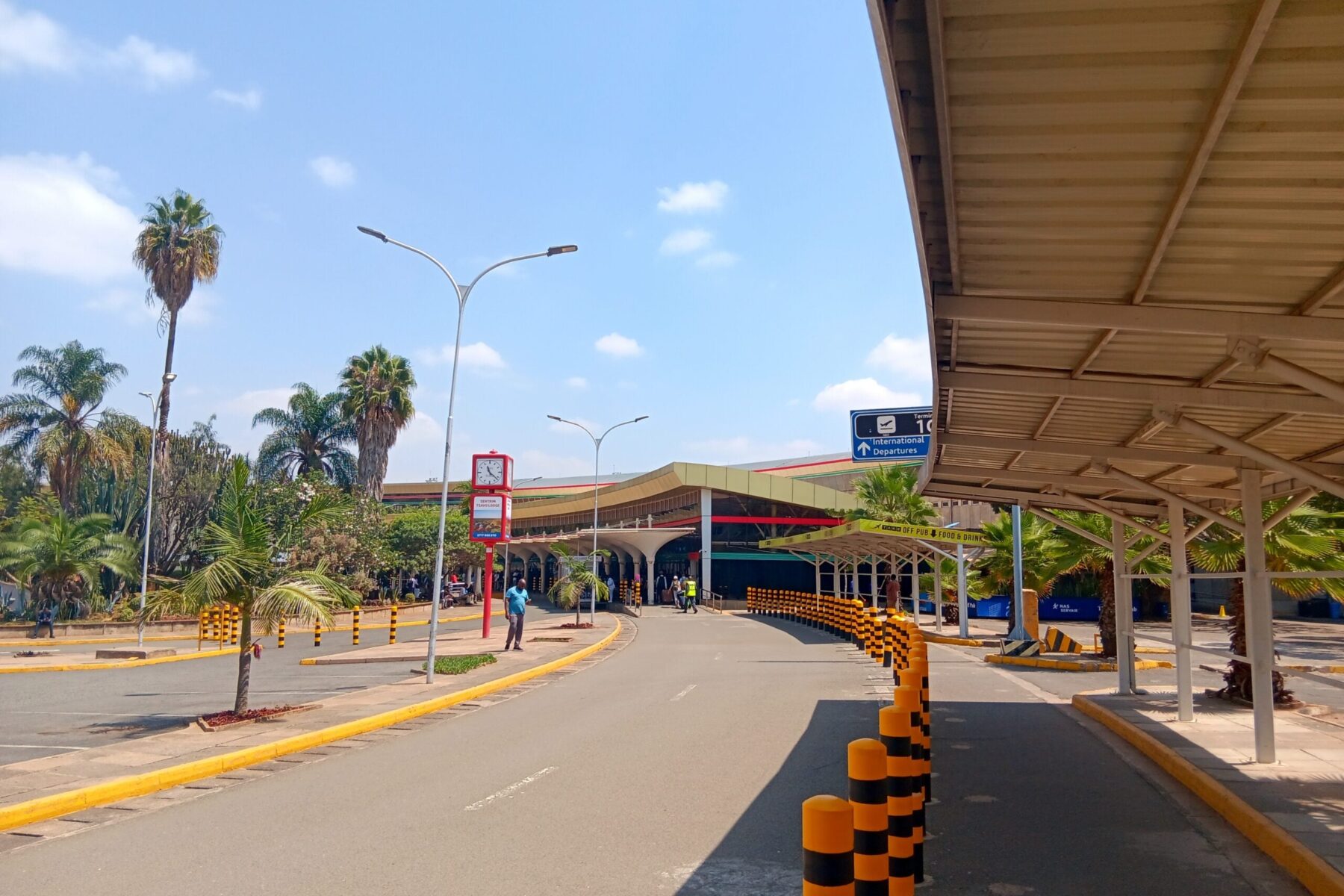 Road leading to the terminal building of Jomo Kenyatta International Airport in Nairobi, with palm trees, black-and-yellow bollards and an International Departures sign