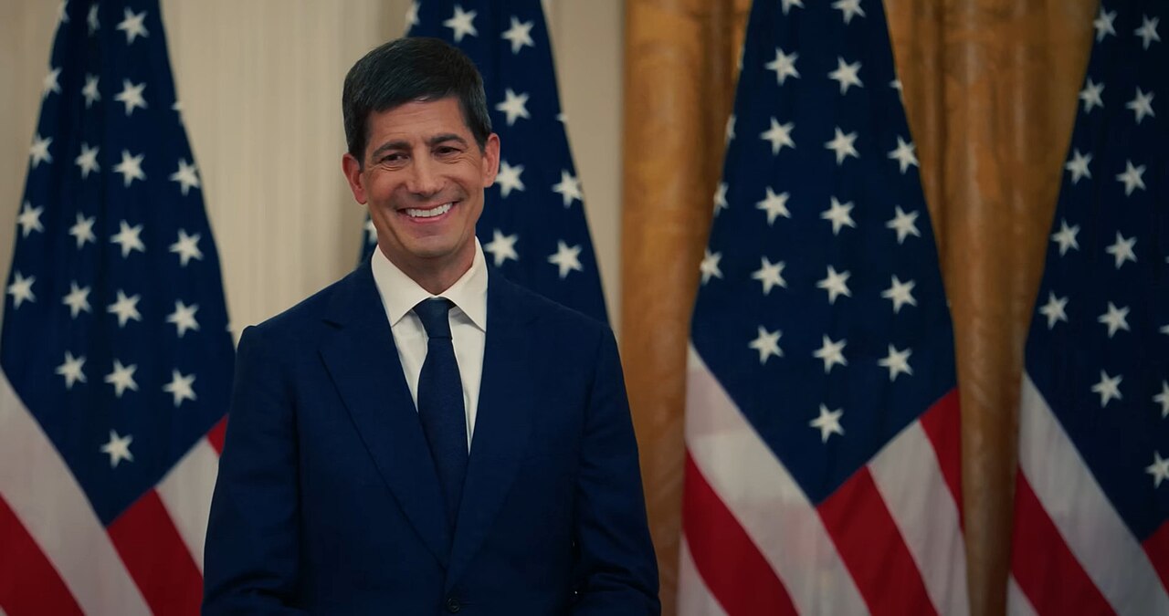 Kevin Warsh, Chair of the US Federal Reserve, smiling in a dark suit in front of American flags