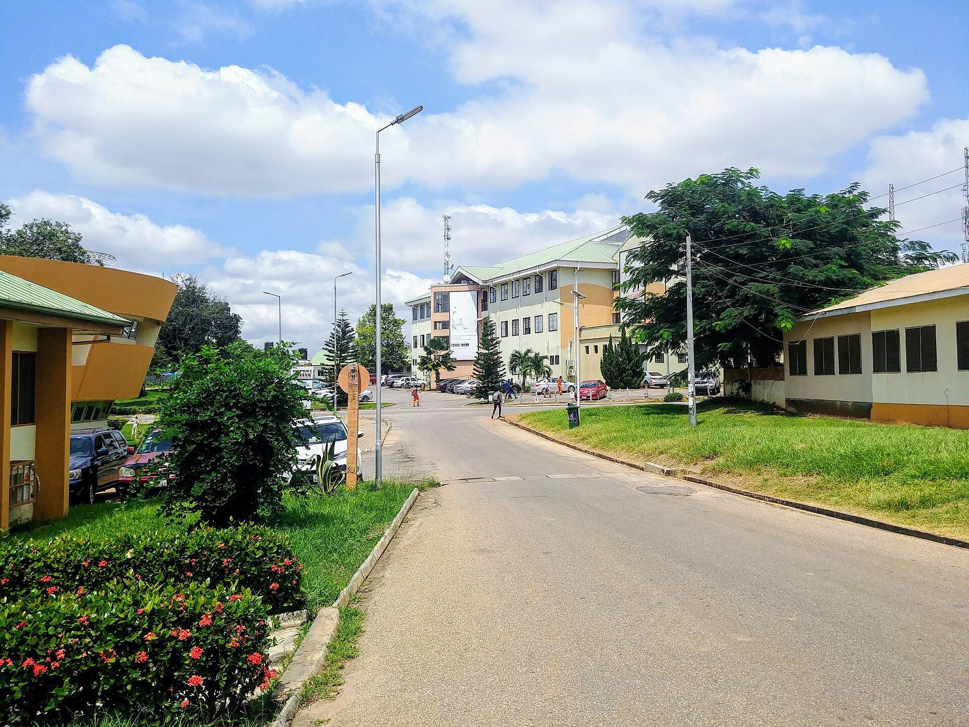 A road through the commercial area of the KNUST campus in Kumasi, Ghana, with parked cars and university buildings