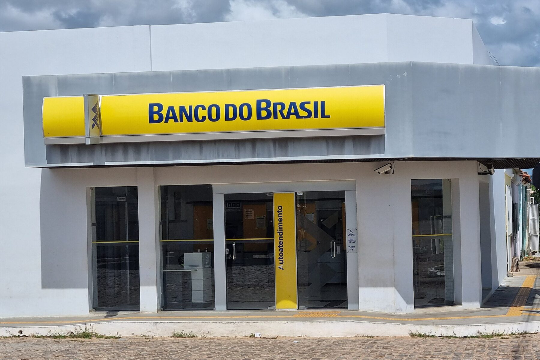 A white Banco do Brasil branch with a yellow sign on a cobbled street under a cloudy sky