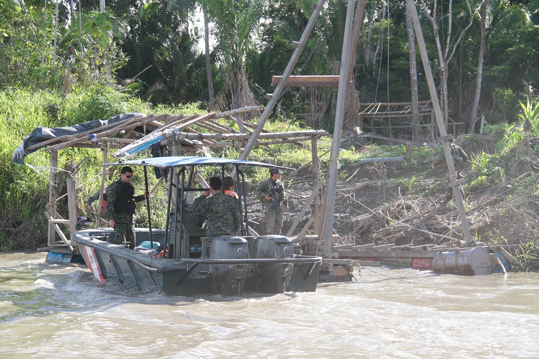 Armed officers in a grey patrol boat beside a wooden illegal gold-mining dredge on a muddy Amazon river