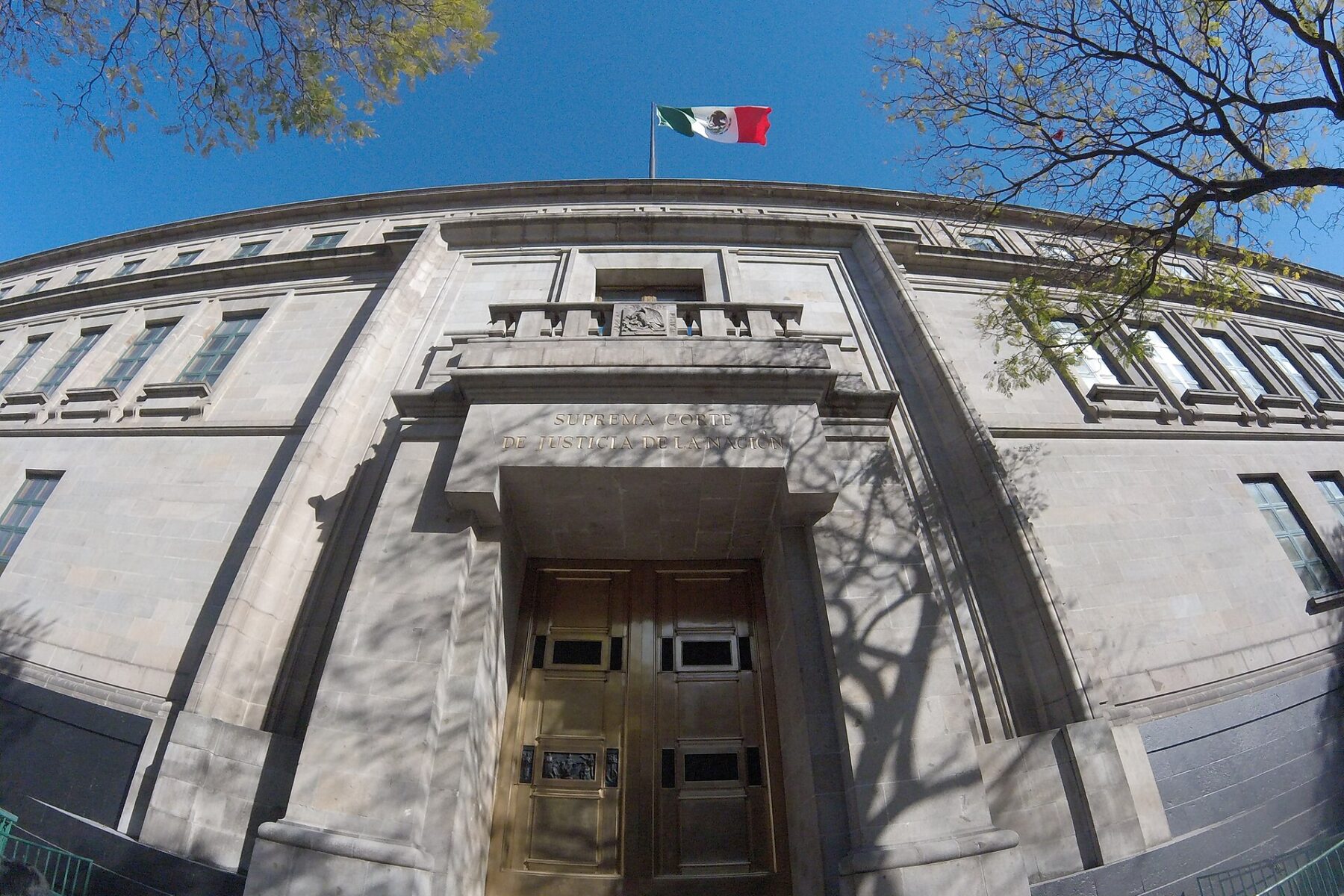The grey stone facade and bronze doors of Mexico's Supreme Court building, with a Mexican flag on top