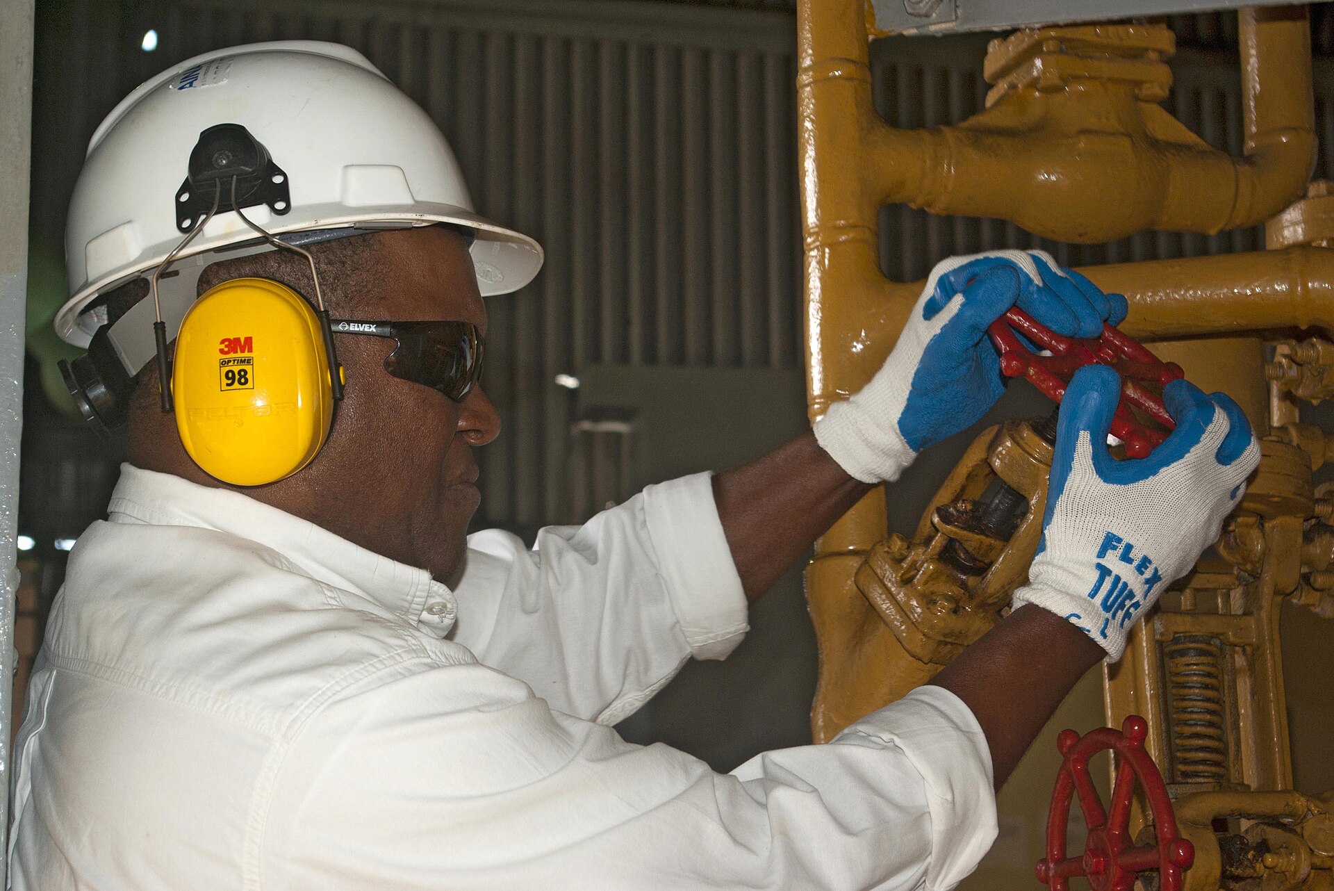 A worker in a white helmet, ear protection and gloves checks yellow valves at a power plant