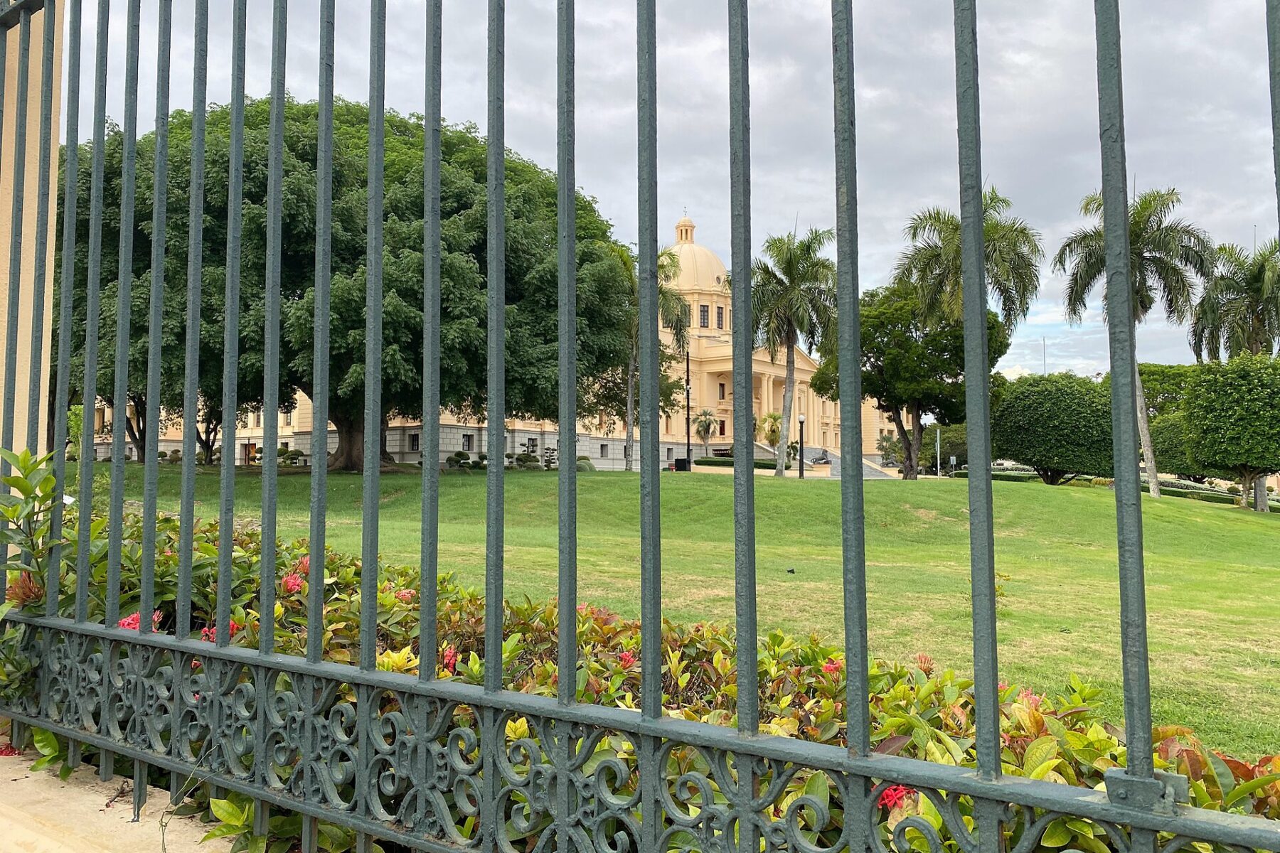 The domed National Palace in Santo Domingo seen through a green iron fence across a lawn with palm trees