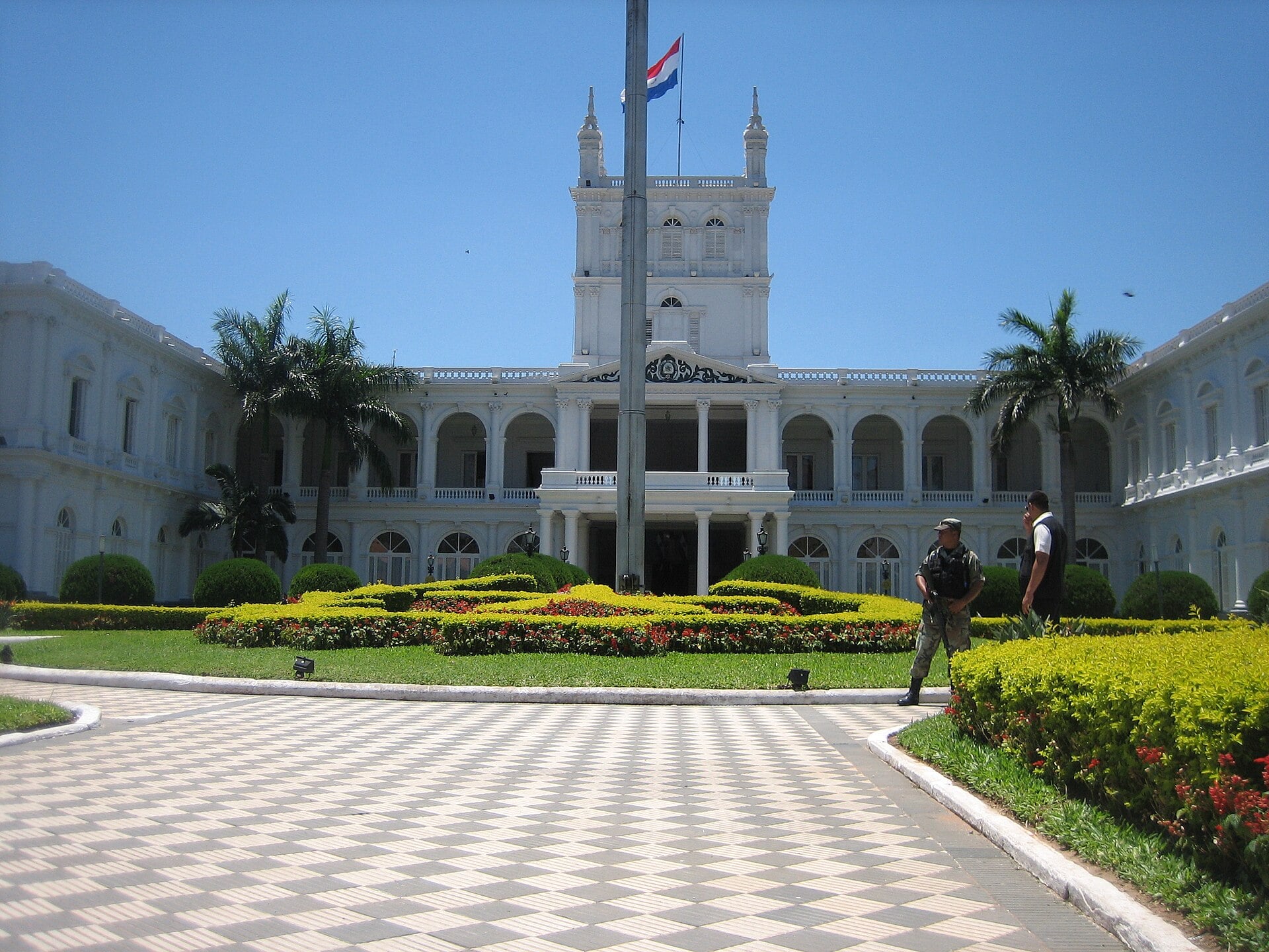 The white Palacio de los López in Asunción with a central tower and Paraguayan flag behind gardens and a checkered plaza