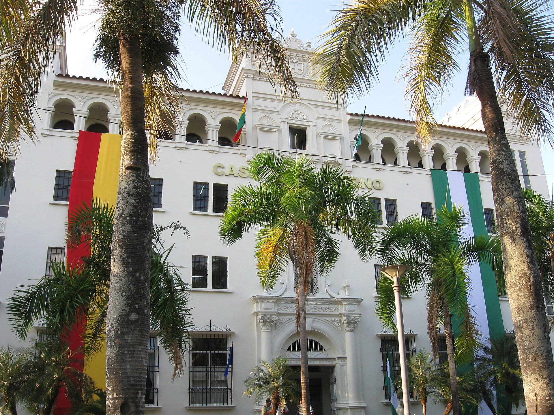 The white historic Casa de Gobierno building in Santa Cruz de la Sierra behind palm trees, with a large green and white flag