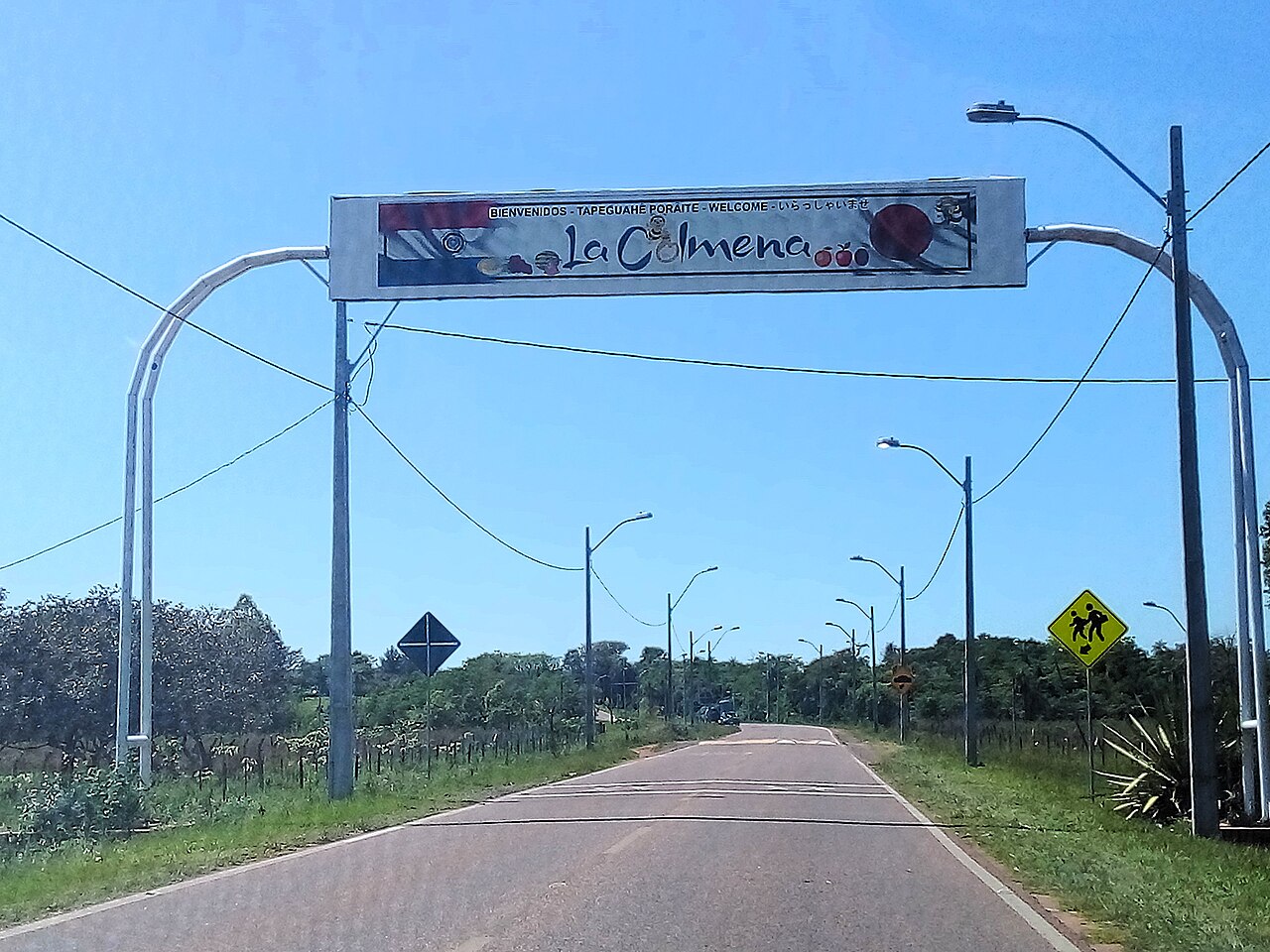 A welcome arch reading La Colmena over a rural road lined with street lamps, trees and a school-crossing sign in Paraguay