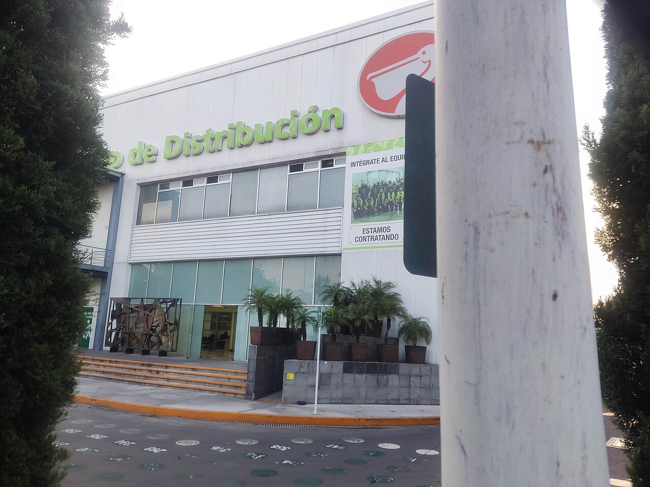 Front of a La Comer distribution centre with the pelican logo, green lettering and a hiring banner