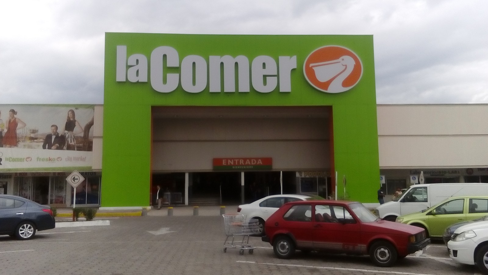 Green entrance facade of a La Comer supermarket with the pelican logo and a car park in front