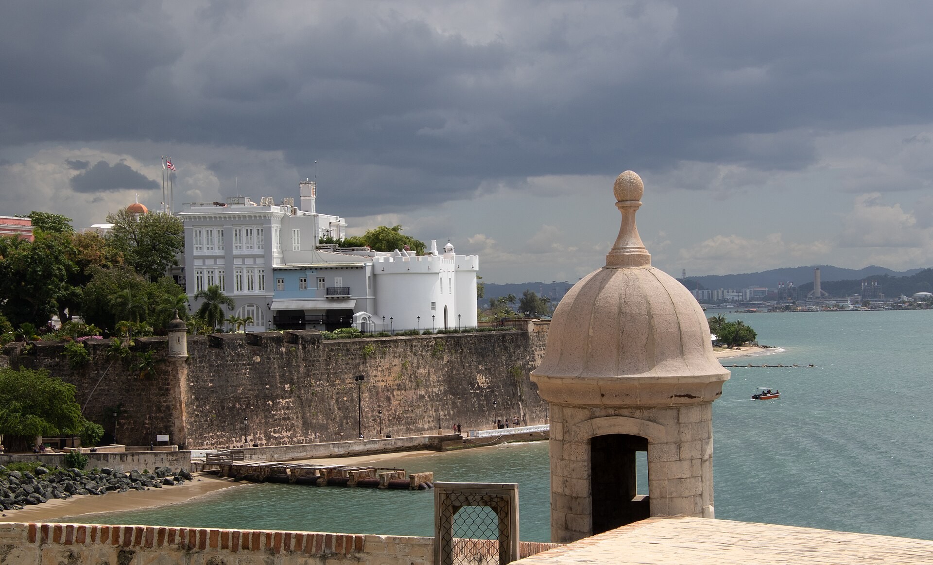 The white La Fortaleza mansion in Old San Juan above the sea wall and harbour, with a stone sentry box in the foreground under a stormy sky