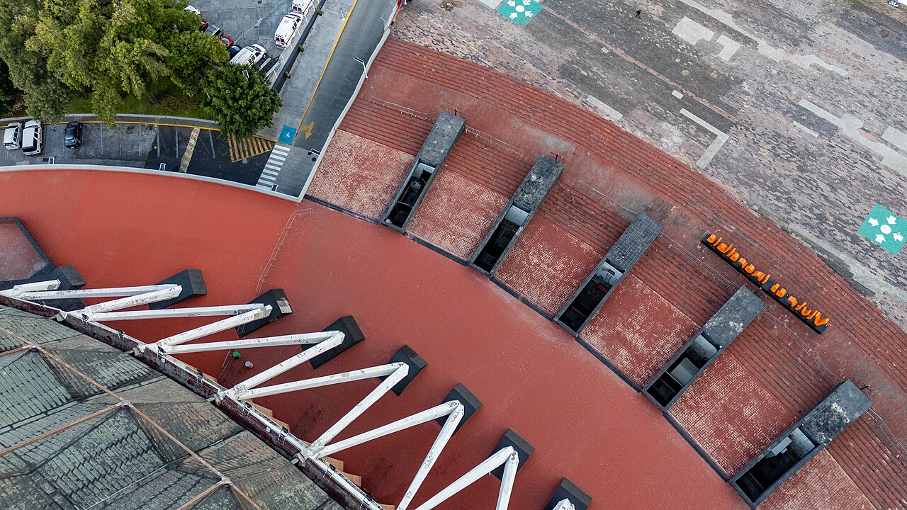 Aerial view of the red brick edge and white roof struts of the Palacio de los Deportes in Mexico City, with orange lettering, a street and parked cars beside it.