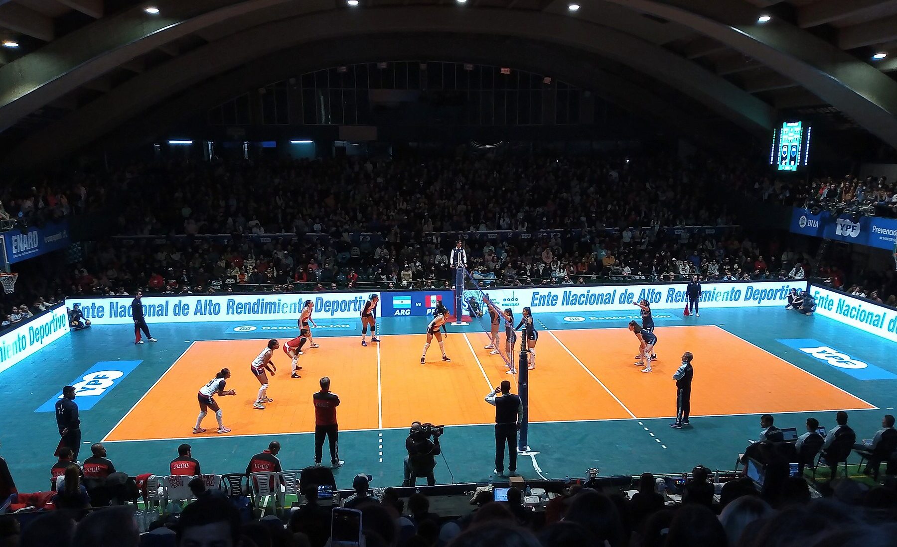 Wide view of an indoor arena during a women's volleyball match on an orange court, with a packed crowd in the stands under a curved roof