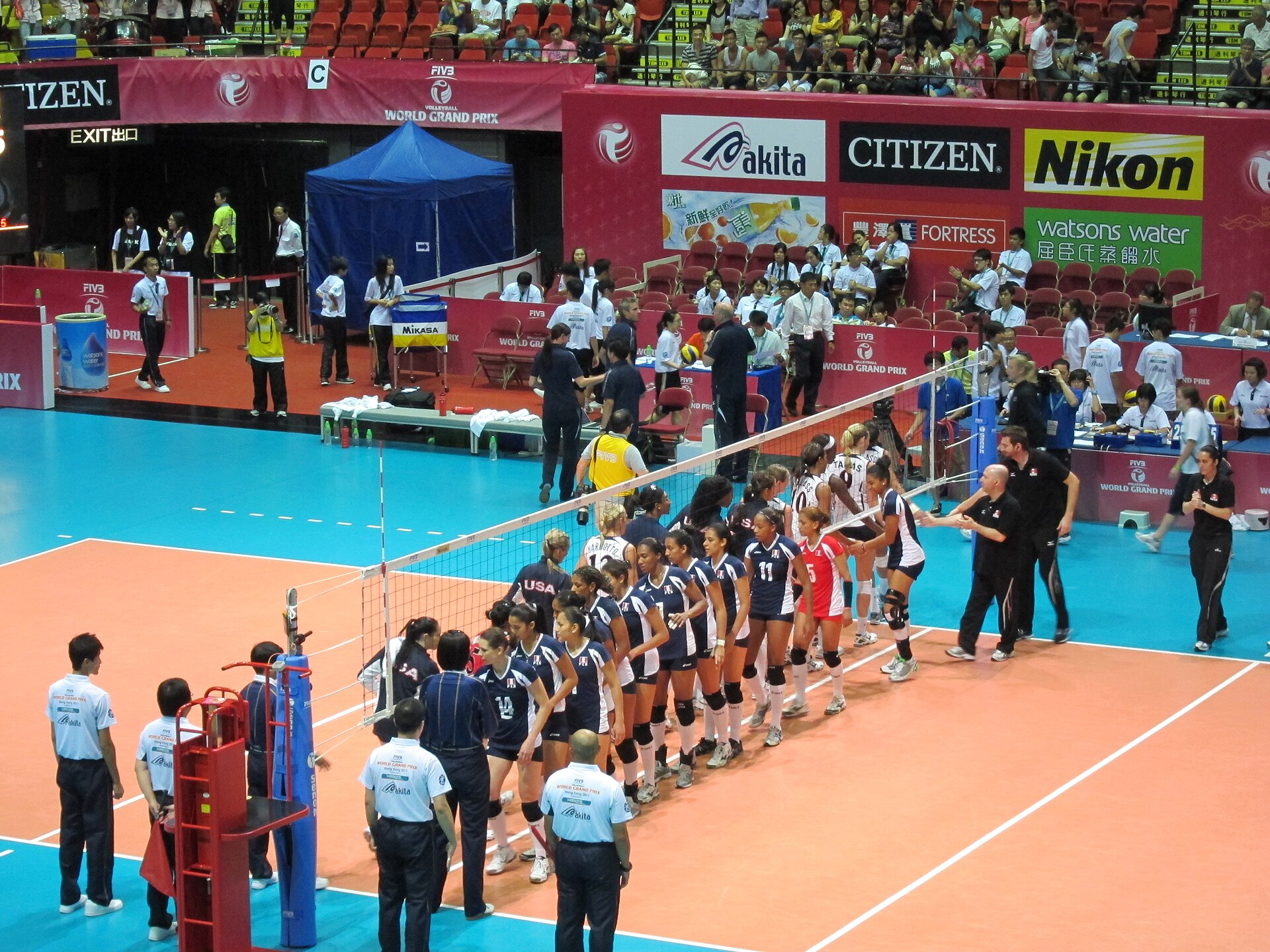Women's volleyball players from Peru in white and red and the United States in navy shake hands along the net in an arena with sponsor boards