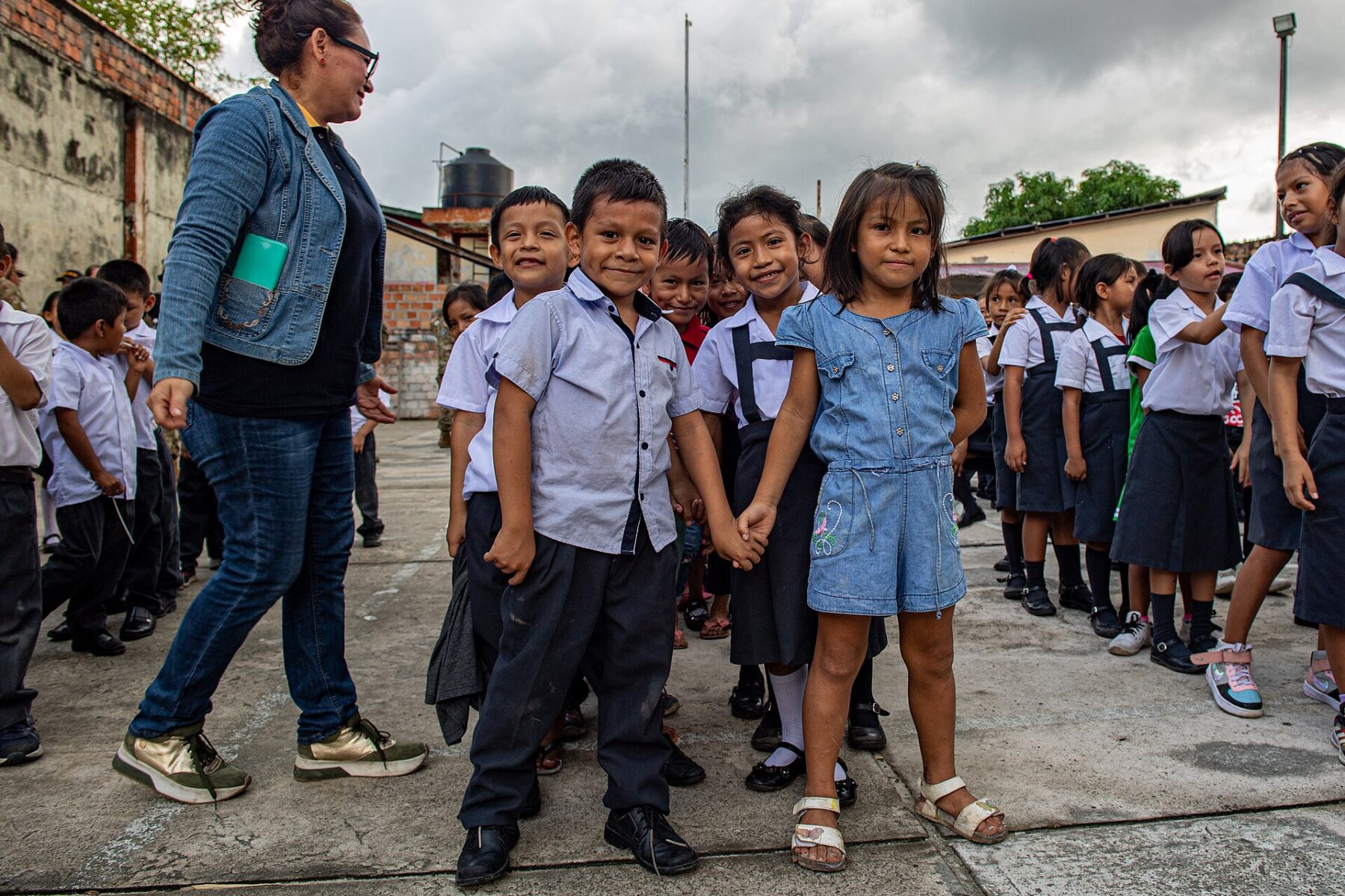 Smiling primary school children in grey and blue uniforms stand in a concrete schoolyard with a teacher in a denim jacket beside them