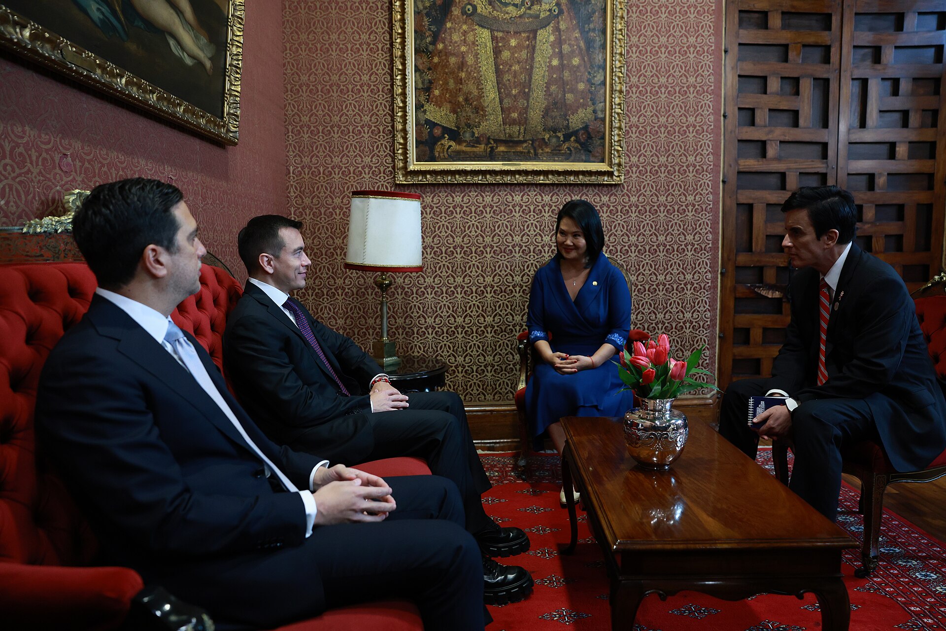 A woman in a blue dress sits in an armchair in an ornate room with red sofas and paintings, talking with three men in suits