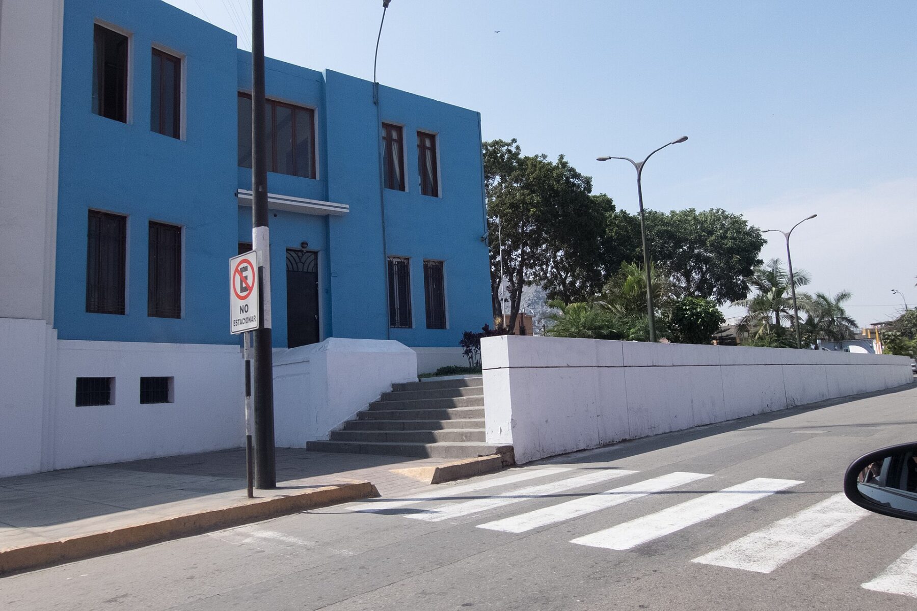A blue two-storey building with barred windows beside a long white wall and a zebra crossing on a sunny street